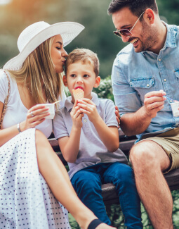 Eltern und kleiner Sohn sitzen lächelnd im Freien und essen gemeinsam Eis