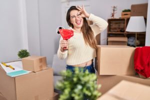 young hispanic woman moving new home packing boxes smiling happy doing ok sign with hand eye looking through fingers